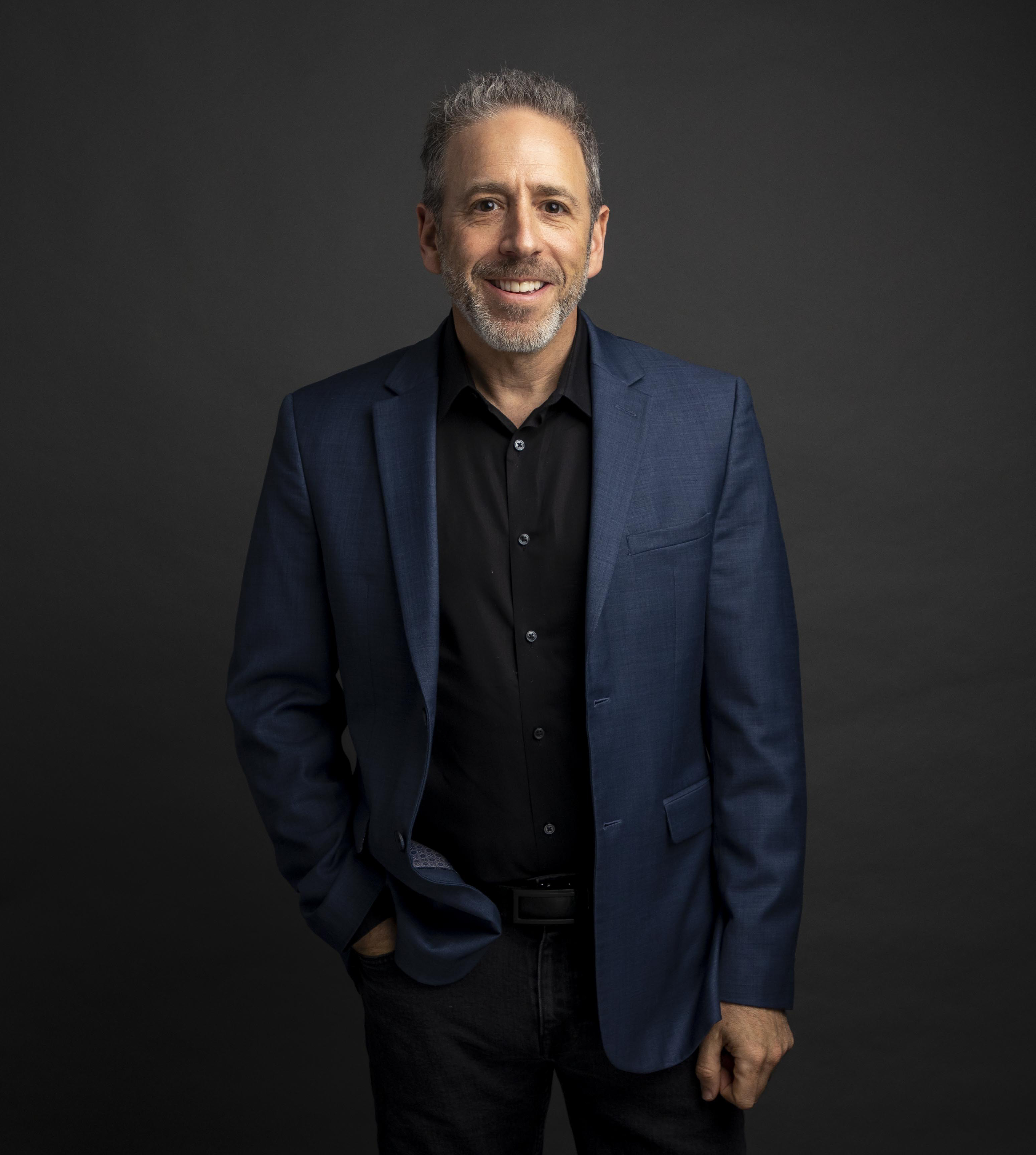 Professional portrait of Alex Heiche, smiling and wearing a blue suit jacket against a dark background.